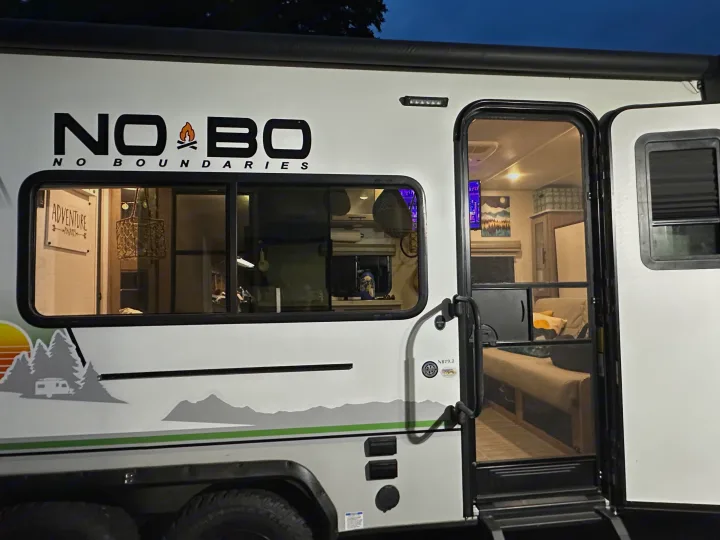 A modern No Boundaries RV rental at dusk, its side graphics visible, with the door open revealing a cozy, lit interior.. No Boundaries 19 2021