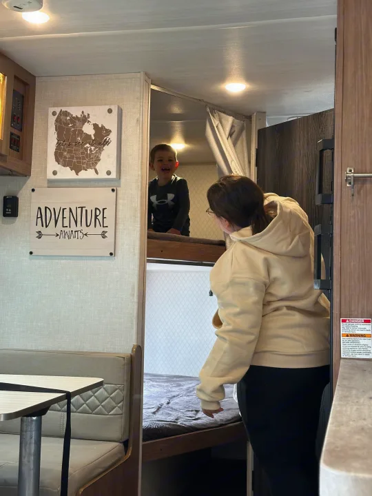 RV rental adventure! A happy child is in a top bunk as an adult looks on. "Adventure Awaits" on the wall inside this cozy camper.. No Boundaries 19 2021