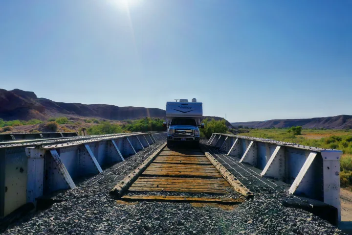 An RV rental truck camper navigates a wooden section of a railroad bridge in a sunlit desert landscape. Ideal for unique travel.. Sportchassis Sportchassis Tow Vehicle 2012