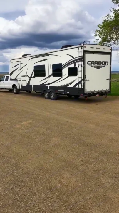 Spacious white and black Carbon toy hauler RV for rent, towed by a pickup truck on a gravel lot.. Sportchassis Sportchassis Tow Vehicle 2012