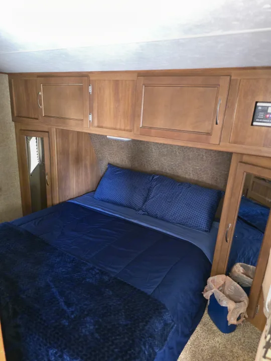 Comfortable RV rental bedroom with blue bed, overhead cabinets, essential controls, and a mirror.. Coachmen Apex 2015