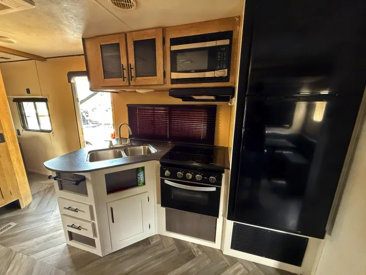 Clean, modern RV rental kitchen with a double sink, full-size fridge, oven, microwave, and stylish light wood cabinetry.. Heartland RVs Prowler 2021