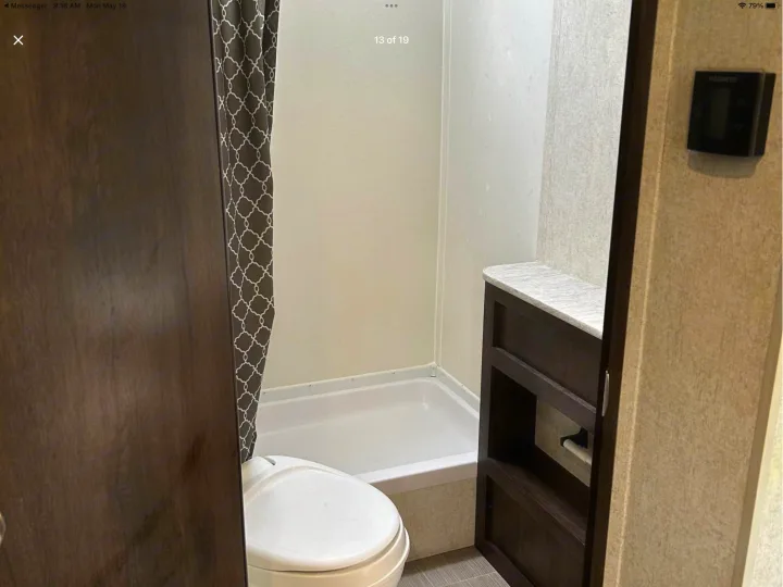 An RV rental bathroom features a white toilet, small shower/tub with patterned curtain, and wooden shelving. A wall control panel is visible.. Starcraft Avalon 2018