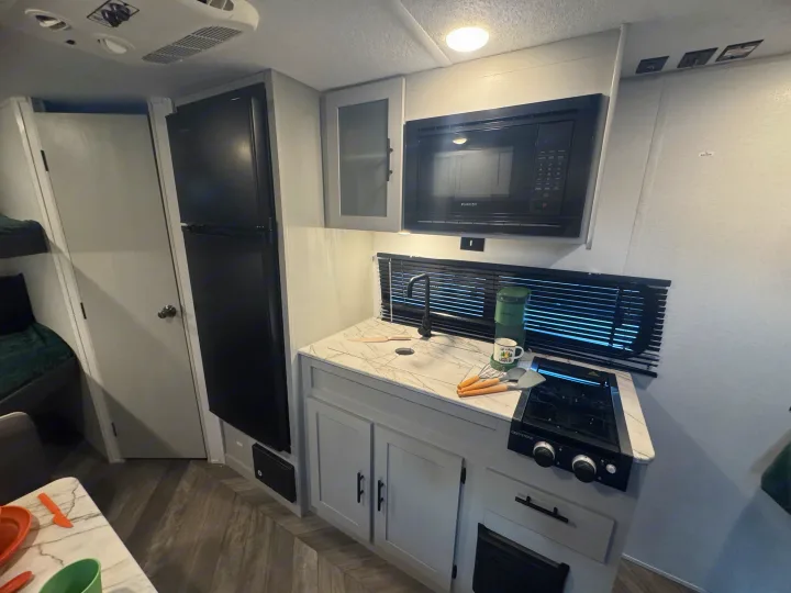 Compact rv rental kitchen with black fridge, microwave, sink, and stove. Light grey cabinets & bunk beds.. Forest River Wildwood FSX 2021