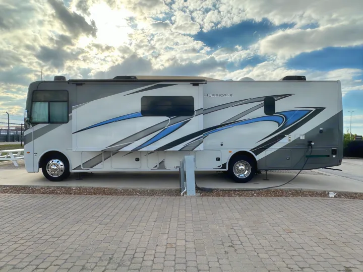 Side view of a white, grey, and blue Hurricane RV rental motorhome parked on pavers with hookups.. Thor Hurricane 2023