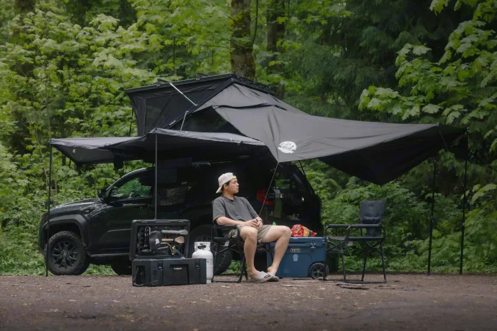 Experience overlanding with an RV rental! Man relaxes under a truck's rooftop tent and awning with camp gear in a forest.. Toyota 4Runner 2025