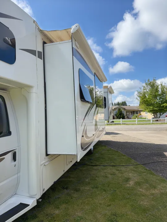 A white RV rental is parked on grass, its slide-out extended with windows visible, under a bright blue sky. Awnings are extended.. Jayco Greyhawk 2015