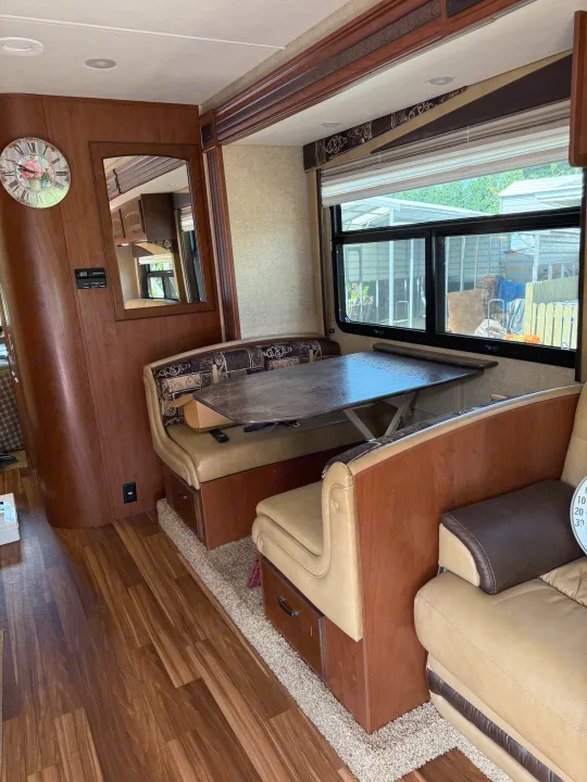 Inside an RV rental: a cozy dinette with tan benches, wood-look flooring, and a window view of outside structures.. Jayco Greyhawk 2015