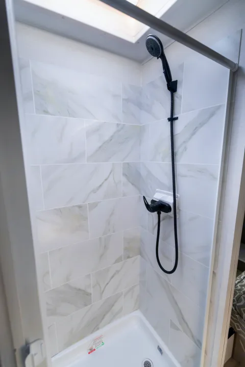Sleek RV rental shower with white marble-look tiles, black fixtures, and a bright overhead skylight, showcasing modern design.. Gulf Stream Conquest 2024