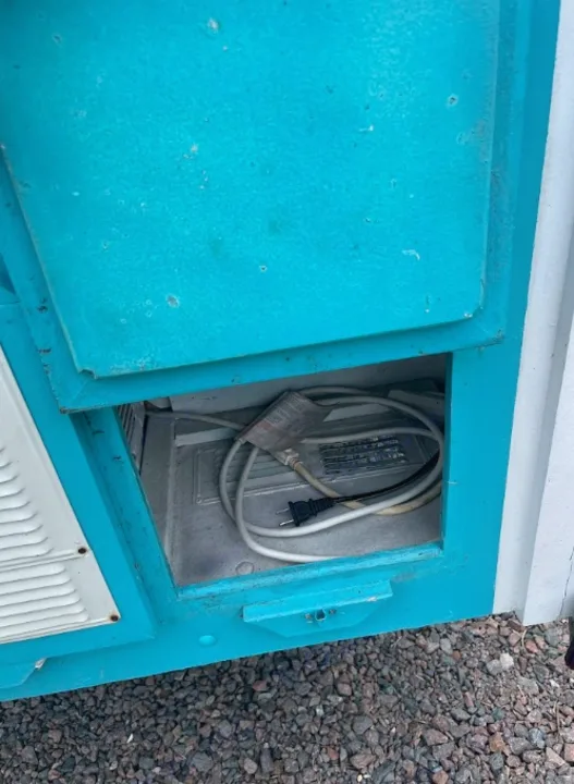 Open electrical compartment on an **rv rental**, revealing power cables and a plug. Teal exterior, gravel below.. YNGQ Box 2017