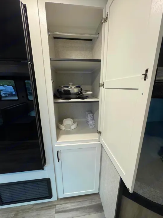 RV rental cabinet showing organized kitchenware: pots, pans, and dishes on shelves, with a lower cabinet and adjacent fridge.. Jayco JAY Flight SLX 261BHS 2025