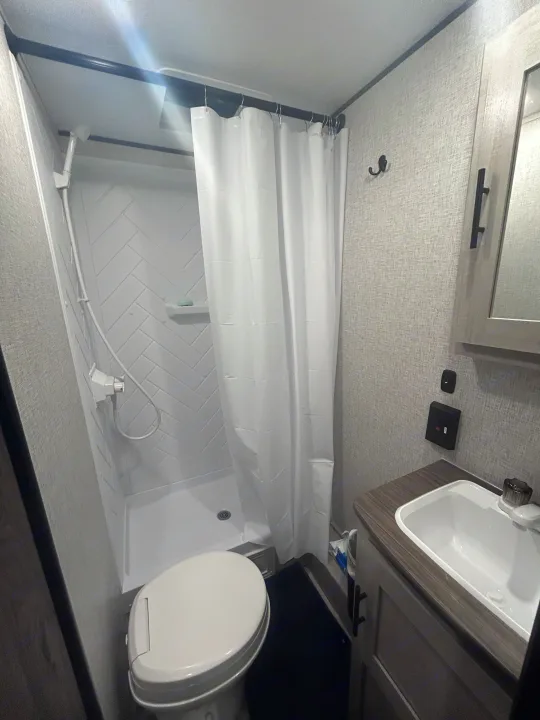 Compact RV rental bathroom with shower, toilet, and sink. White shower curtain and herringbone tile.. Crossroads Zinger Lite 2025