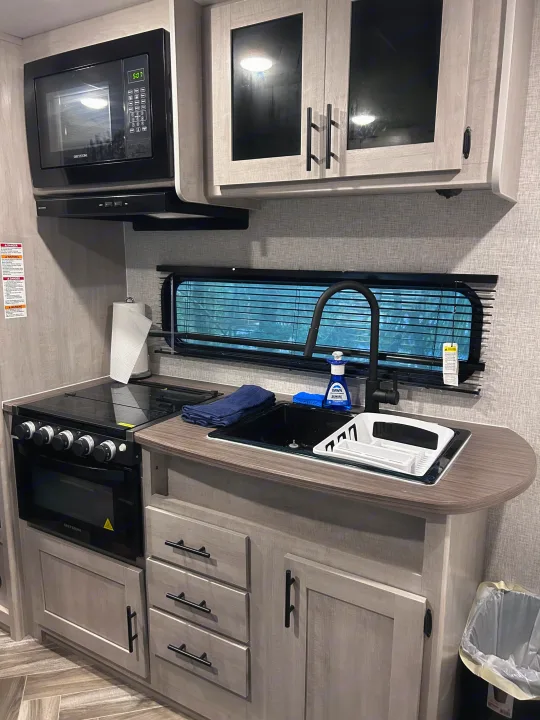RV rental kitchen area: Black microwave, stove, and faucet contrast with light gray cabinets and textured walls, showing a sink.. Crossroads Zinger Lite 2025