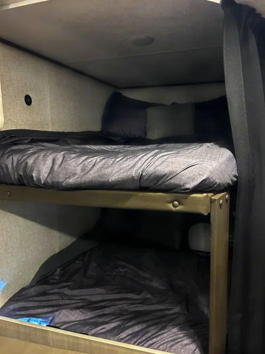 Cozy grey bunk beds in an RV rental with bedding, pillows, and a wall outlet for charging. Dark curtains offer privacy.. Crossroads Zinger Lite 2025