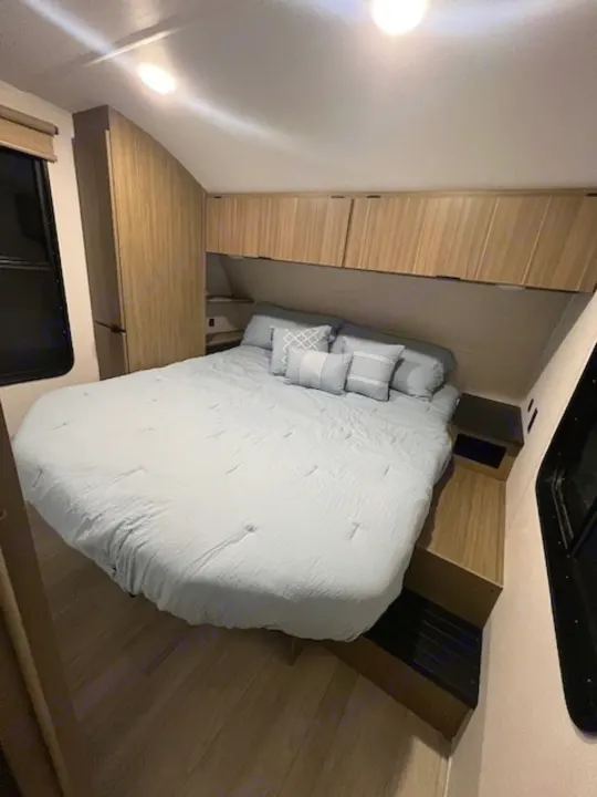 Inside an RV rental, this modern bedroom offers a light blue queen bed, built-in storage, and bright interior lighting.. Forest River 273QBXL 2025
