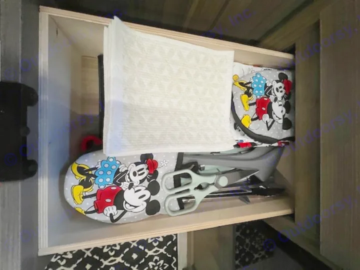 RV rental drawer: Mickey Mouse ironing board, towels, and kitchen tools show smart storage solutions for travel.. Forest River 273QBXL 2025