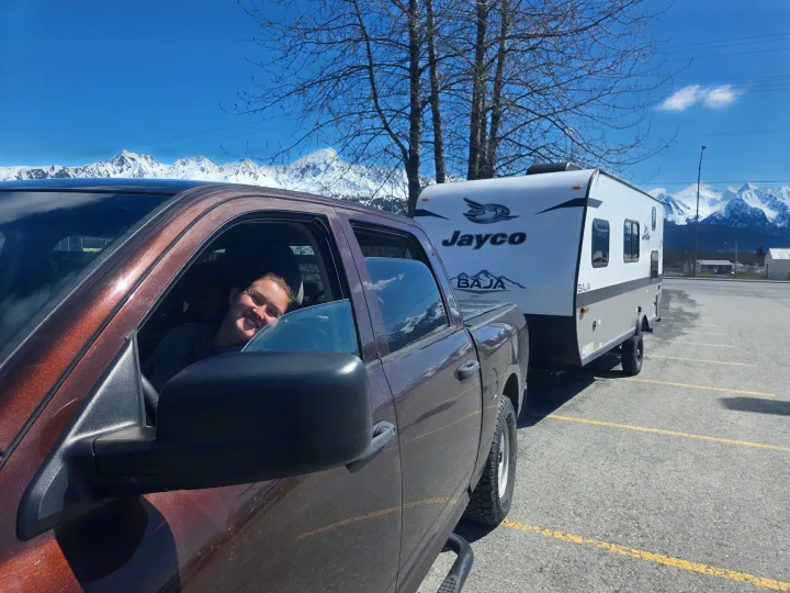 A happy driver in a truck towing a white Jayco Baja RV rental, parked with snow-capped mountains in the background.. Jayco Jay Flight SLX Baja Edition 2022