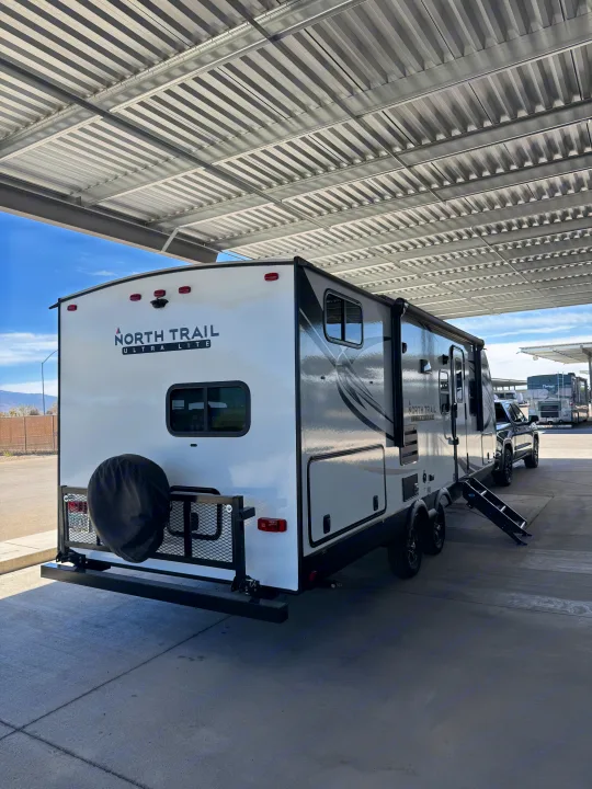 A white North Trail travel trailer, potentially an rv rental, parked under a shelter. Features a rear spare tire and entry steps.. Heartland RVs North Trail 2021
