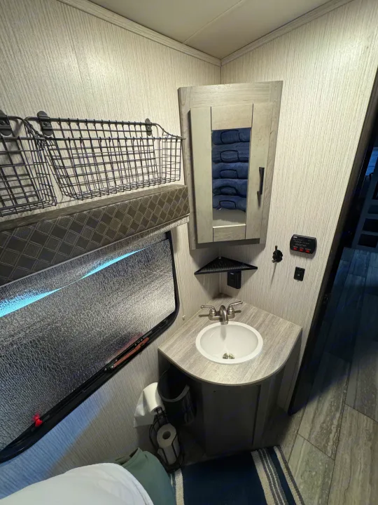 Compact RV rental bathroom featuring a corner sink, mirrored cabinet, and wire wall storage. Window is covered.. Heartland RVs North Trail 2021