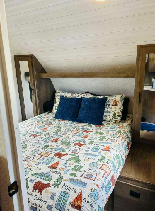 Inside an RV rental: a compact bedroom with a camping-themed bedspread, blue pillows, and wood cabinetry.. Forest River Salem Cruise Lite 2022
