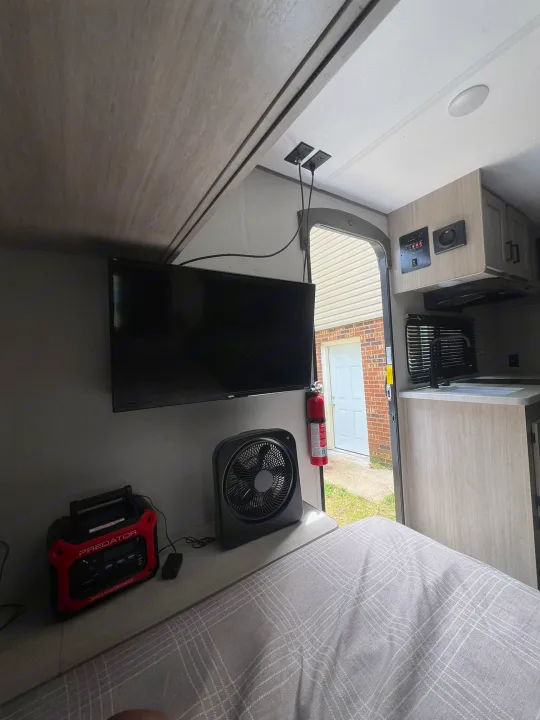 Inside a compact RV rental: bed with plaid blanket, TV, power station, fan, and mini kitchen near the exit.. Forest River Aurora Aurora 2025