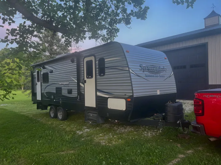 A gray Springdale travel trailer, ideal for an rv rental, is hitched to a red truck, parked near a large barn.. Keystone RV Springdale 2019