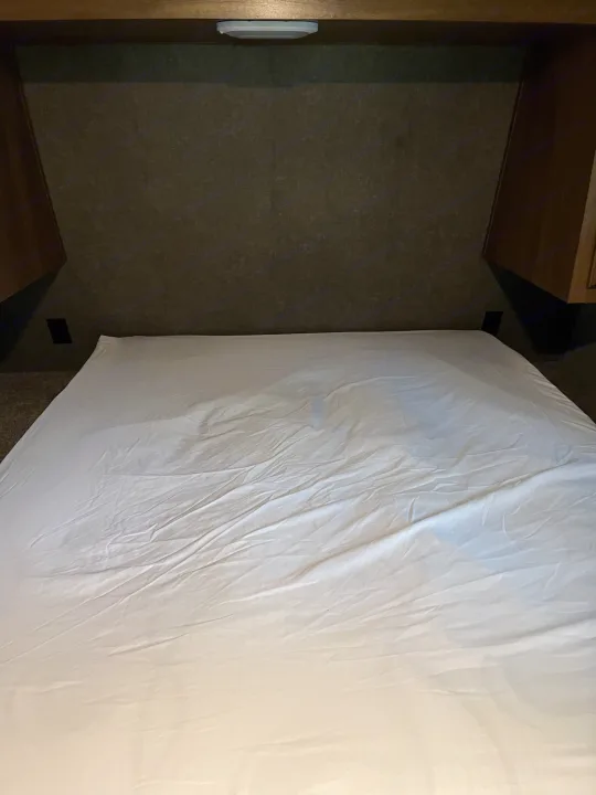 Inside an RV rental, a compact bedroom with a bed, white sheets, and built-in amenities like outlets and an overhead light.. Keystone RV Springdale 2019