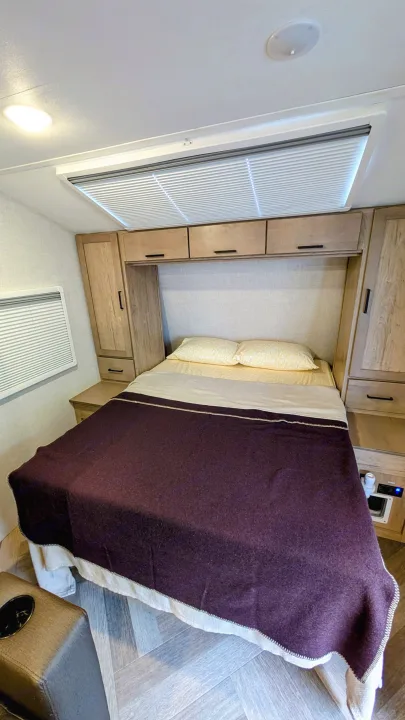 Compact RV rental bedroom: bed with purple blanket, wood cabinets, window with blinds, and ceiling lights. Ideal for travel.. Ember Overland 2023