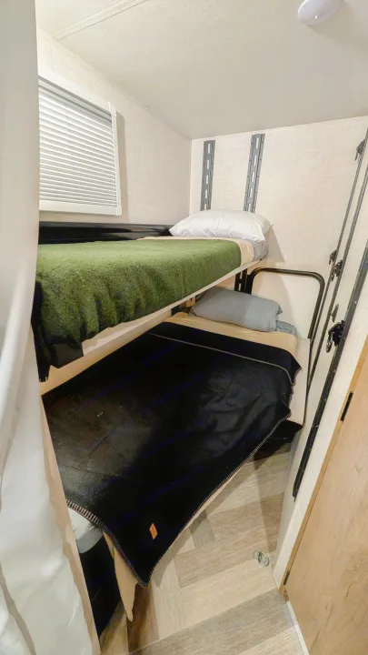 Cozy bunk beds inside an RV rental feature green and black blankets, a window, and wall tracks on wood-look flooring.. Ember Overland 2023