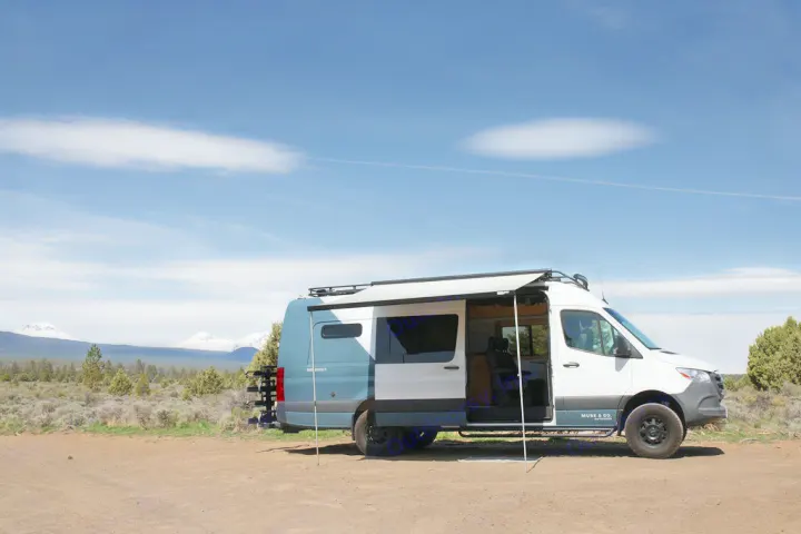 Modern white & blue Sprinter camper van, an ideal RV rental, parked on a dirt road, snow-capped mountains in background. Awning extended.. Mercedes-Benz Sprinter 2020