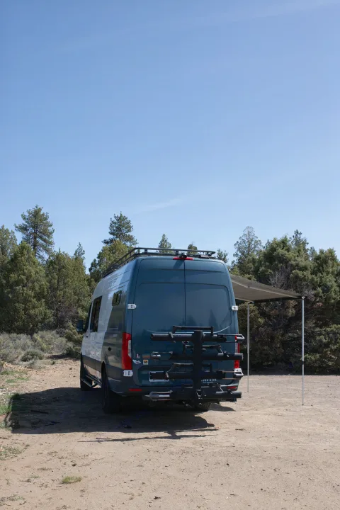Dark Sprinter RV rental camper van on a dirt road amidst trees, with an extended side awning and rear bike rack under clear skies.. Mercedes-Benz Sprinter 2020
