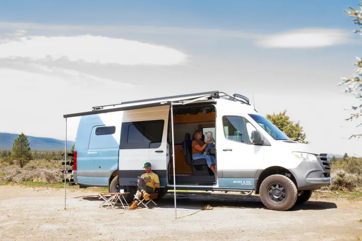 A family relaxes in an RV rental camper van in a scenic outdoor setting. A man reads outside, a woman holds a baby inside.. Mercedes-Benz Sprinter 2020