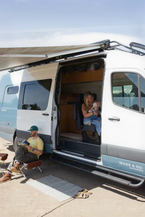 Family enjoying their RV rental: A man reads outdoors while a woman holds a baby inside their camper van.. Mercedes-Benz Sprinter 2020