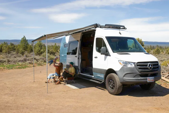 A family with a baby enjoys a relaxing day outside their white Mercedes Sprinter RV rental, parked with an awning extended in nature.. Mercedes-Benz Sprinter 2020