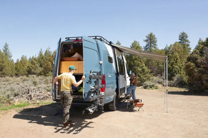 RV rental adventure: A family sets up their van with an awning extended, bikes on back, for camping in nature.. Mercedes-Benz Sprinter 2020