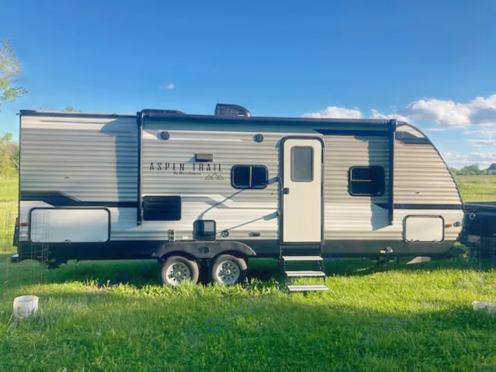 This grey and white Aspen Trail travel trailer is ready for your RV rental adventure in a sunny field.. Dutchmen 2340BHS 2020