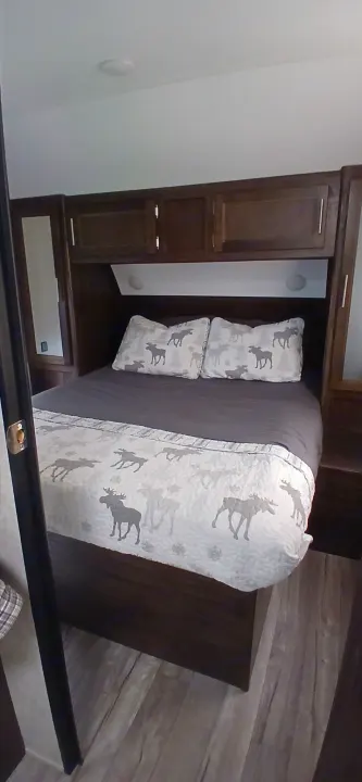 This RV rental bedroom features a cozy bed with moose-patterned bedding, dark wood cabinets, and overhead lighting.. Forest River Cherokee Grey Wolf 2019