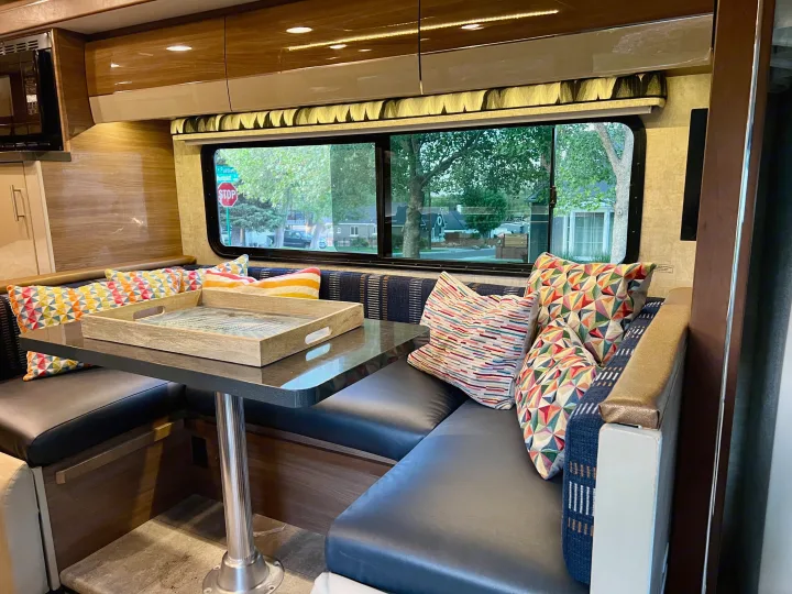 Inside a spacious RV rental, a cozy dinette with dark seating, a table, and colorful pillows. Large windows show the street.. Itasca Navion 2015