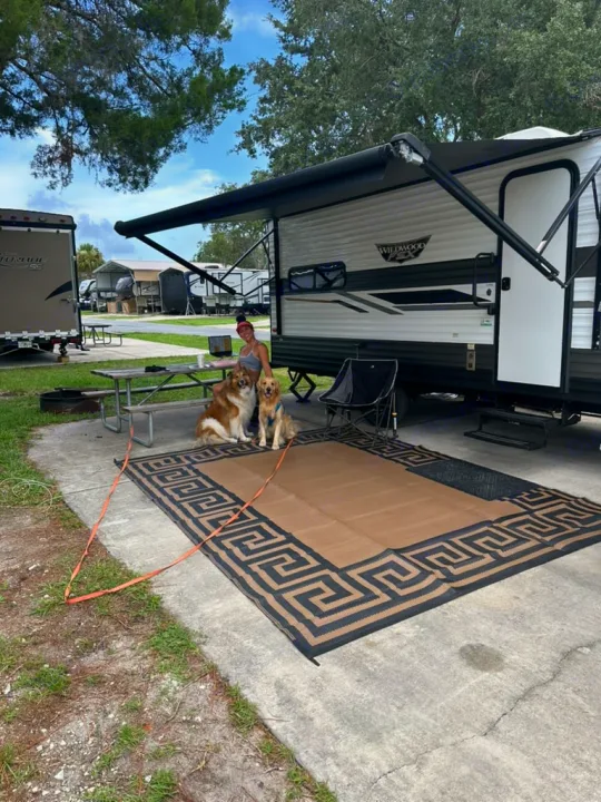 Relaxing RV rental setup: Woman with two dogs at a spacious campsite, featuring an RV with an awning and large outdoor mat.. Forest River Wildwood FSX 2022