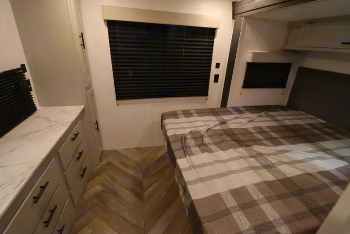 Modern RV rental bedroom: plaid bed, dresser w/ marble top, and herringbone floor. Compact comfort for your travels.. Forest River Wildwood FSX 2022