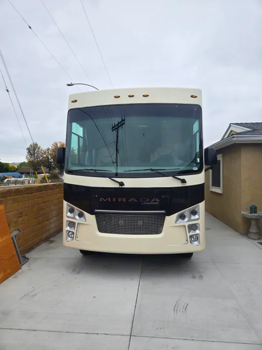 A cream-colored Coachmen Mirada RV rental, parked head-on in a driveway with a house nearby.. Coachmen Mirada 2020