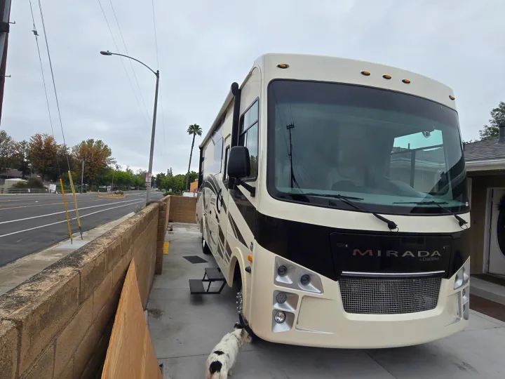 A large, white and black Mirada motorhome is parked on a driveway with a small dog by its wheel, ready for an RV rental.. Coachmen Mirada 2020