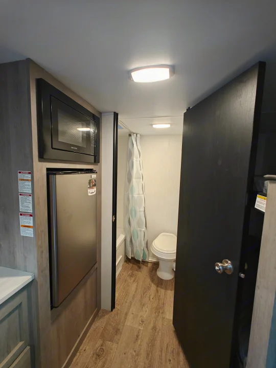 RV rental interior: Left shows kitchen with microwave & fridge. Right features an open bathroom with toilet & shower. Wood-look floor.. Cruiser Avenir 17BH Travel Trailer Avenir 17BH 2025