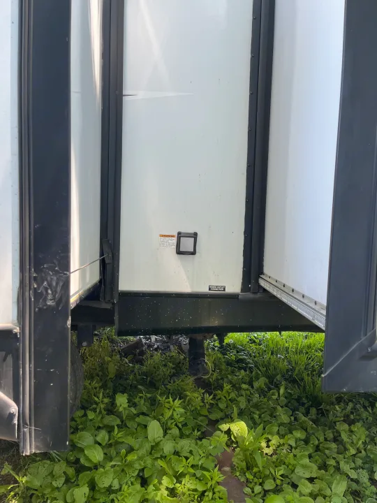 Side of an RV rental with its slide-out partially extended. A sewer outlet connection is visible near the bottom, surrounded by green foliage.. Forest River Tracer Breeze 2019