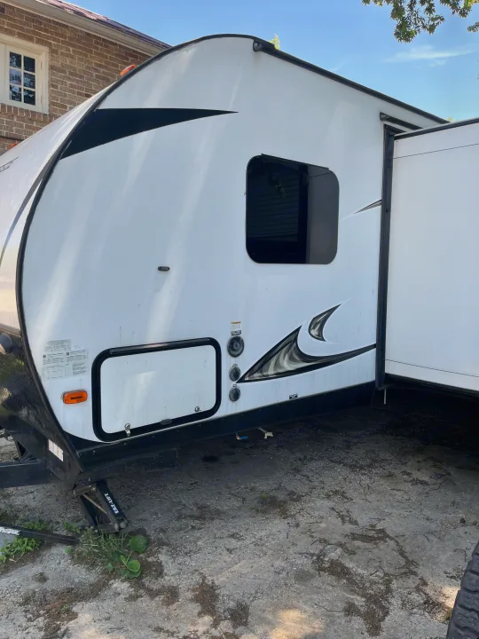 Exterior side view of a white and black RV rental, revealing water hookups, a side window, and an open door, parked on gravel.. Forest River Tracer Breeze 2019