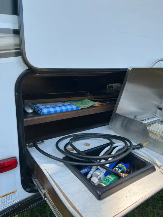 An RV rental's pull-out outdoor cooking area with a grill, sink, propane hose, and camping supplies.. Forest River Tracer Breeze 2019