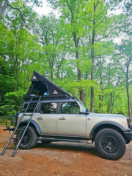 Ford Bronco SUV with rooftop tent & ladder for camping, parked in a forest. Ideal for outdoor adventures, like a compact RV rental.. Ford Bronco Outer Banks 2022