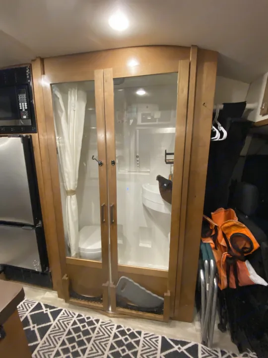 Inside an RV rental, a compact bathroom with toilet, sink, and shower is visible through glass doors, next to kitchen appliances.. Forest River Coachmen Crossfit 2018