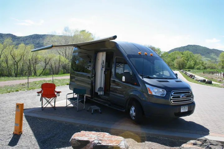 Gray Ford Transit RV rental camper van with extended awning & chairs at a scenic mountain campsite, ready for outdoor travel.. Forest River Coachmen Crossfit 2018