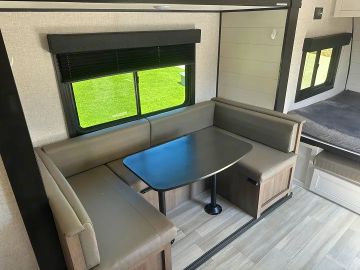 This RV rental interior features a spacious U-shaped dinette with grey cushions, a dark table, and windows showing green grass. A bed is visible in the background.. Autumn Ridge 188BHS 2025